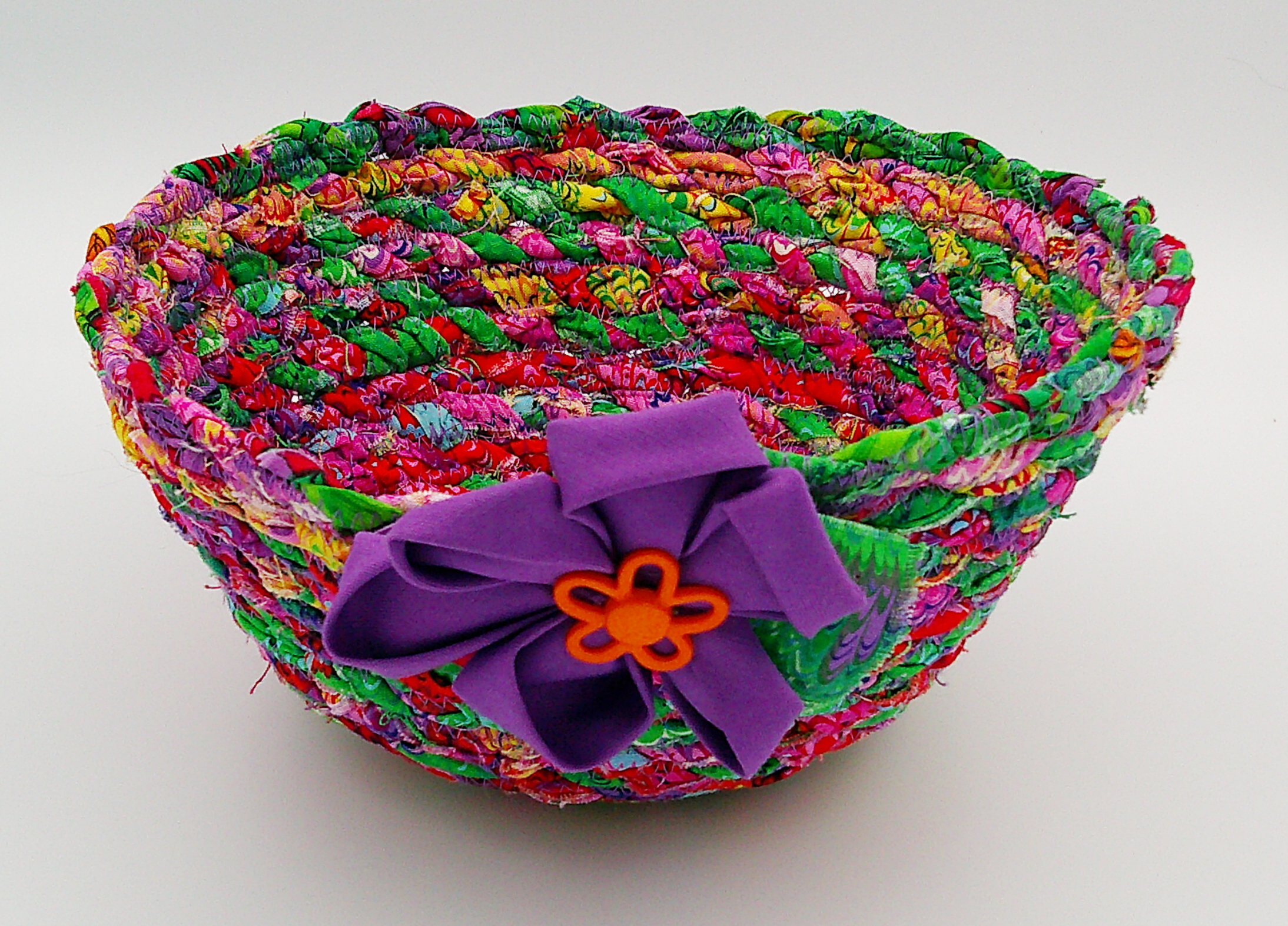 This vibrant multi-colored fabric rope bowl is ready for multiple uses. This view shows the purple flower with orange button. This vibrant multi-colored fabric rope bowl is ready for multiple uses. This view shows the purple flower with orange button.